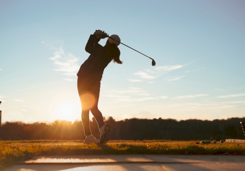 A person is silhouetted against the sunset, swinging a golf club on a grassy field, suggesting an evening round of golf.