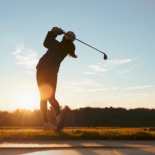 A person is silhouetted against the sunset, swinging a golf club on a grassy field, suggesting an evening round of golf.