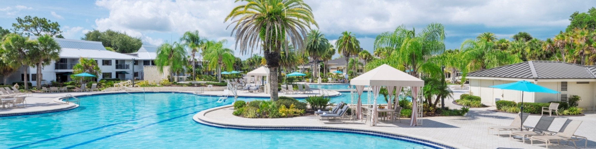 A serene swimming pool with clear blue water, surrounded by palm trees and lounge chairs under white canopies on a partly cloudy day.