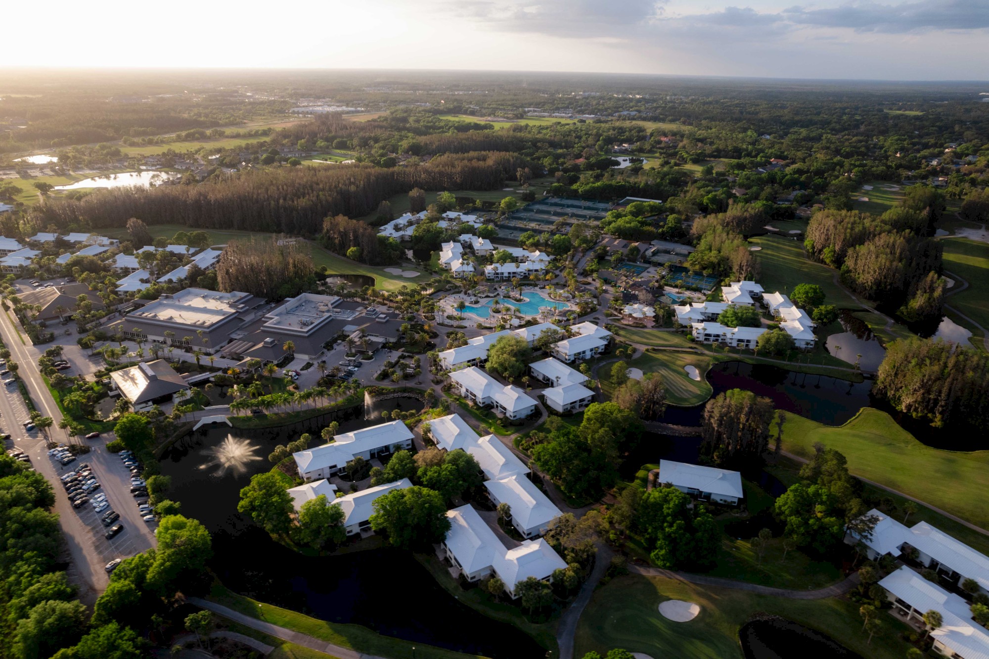 An aerial view of a resort complex with buildings, pools, tennis courts, and surrounding greenery under a partly cloudy sky.