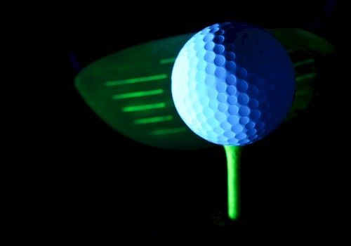 A glowing golf ball on a neon tee in front of a golf club head, set against a dark background.