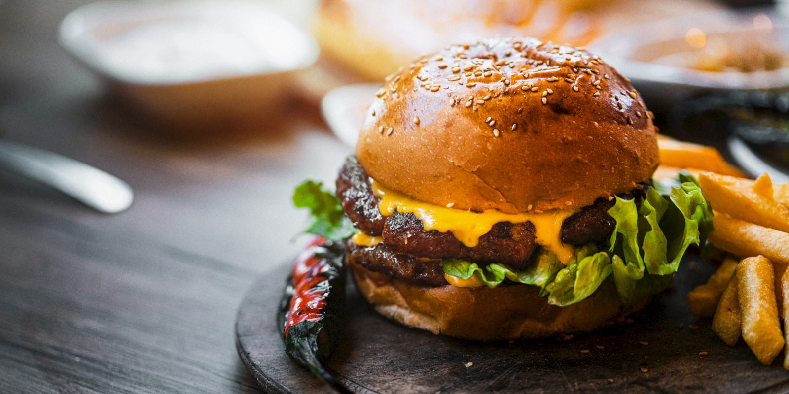 A cheeseburger with lettuce and tomato on a wooden plate, accompanied by fries and a chili pepper, sits on a rustic table.