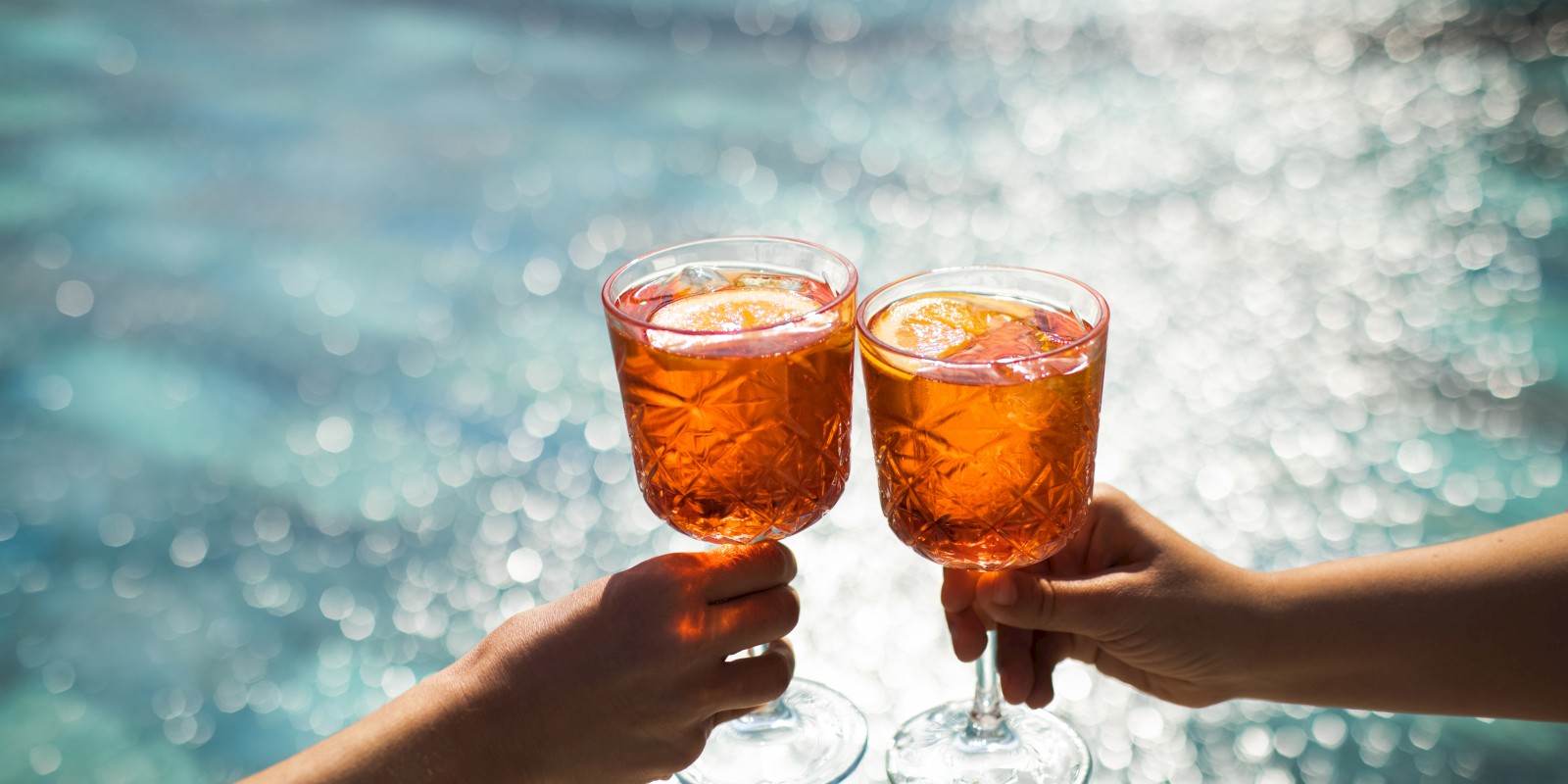 Two hands clinking glasses filled with an orange drink, against a sparkling water background.