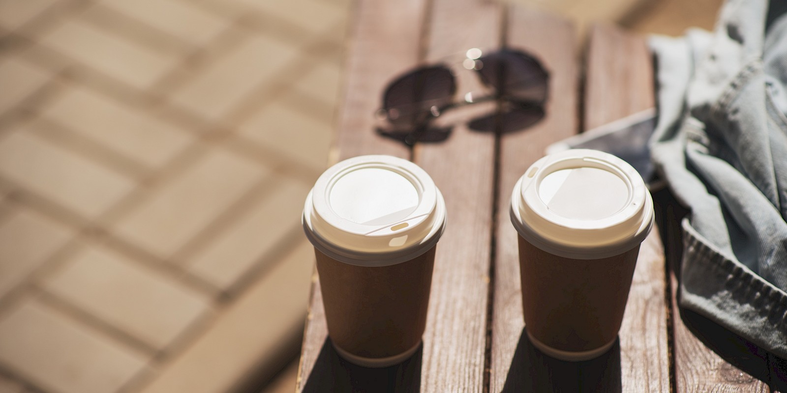 Two takeaway coffee cups sit on a wooden bench with sunglasses and a denim jacket beside them in sunny outdoor setting.